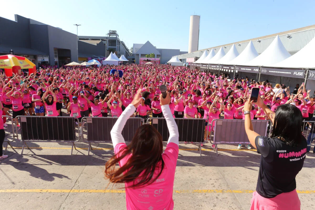Grande grupo de participantes usando camisetas rosa e levantando os braços durante um evento esportivo ao ar livre, diante de um palco e de tendas brancas.